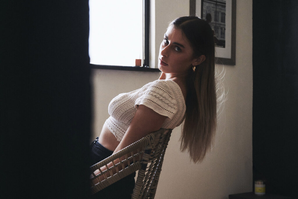 Marintia Bustos a model with long brown hair is seated on a wicker chair looking over her shoulder at the camera. She wears a white lace top and has a framed photograph on the wall behind her. The room has a minimalist design with a window letting in natural light.